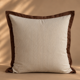 A square throw pillow with a cream and brown vertically striped ribbed fabric body and a solid dark brown border, set against a warm beige background with soft leaf shadow overlays.