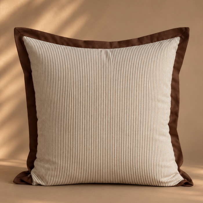 A square throw pillow with a cream and brown vertically striped ribbed fabric body and a solid dark brown border, set against a warm beige background with soft leaf shadow overlays.