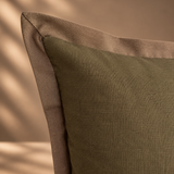 A close-up of a textured olive green throw pillow with a contrasting taupe-brown border, set against a warm beige background with soft leaf shadows.