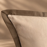 A close-up of a decorative throw pillow with a light beige linen-textured center and a contrasting dark taupe piped border, set against a warm beige background with soft leaf shadows.