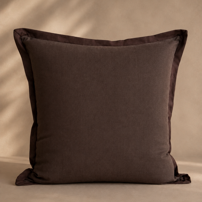 A square dark brown throw pillow with a satin border, displayed against a warm beige background with soft leaf shadows.